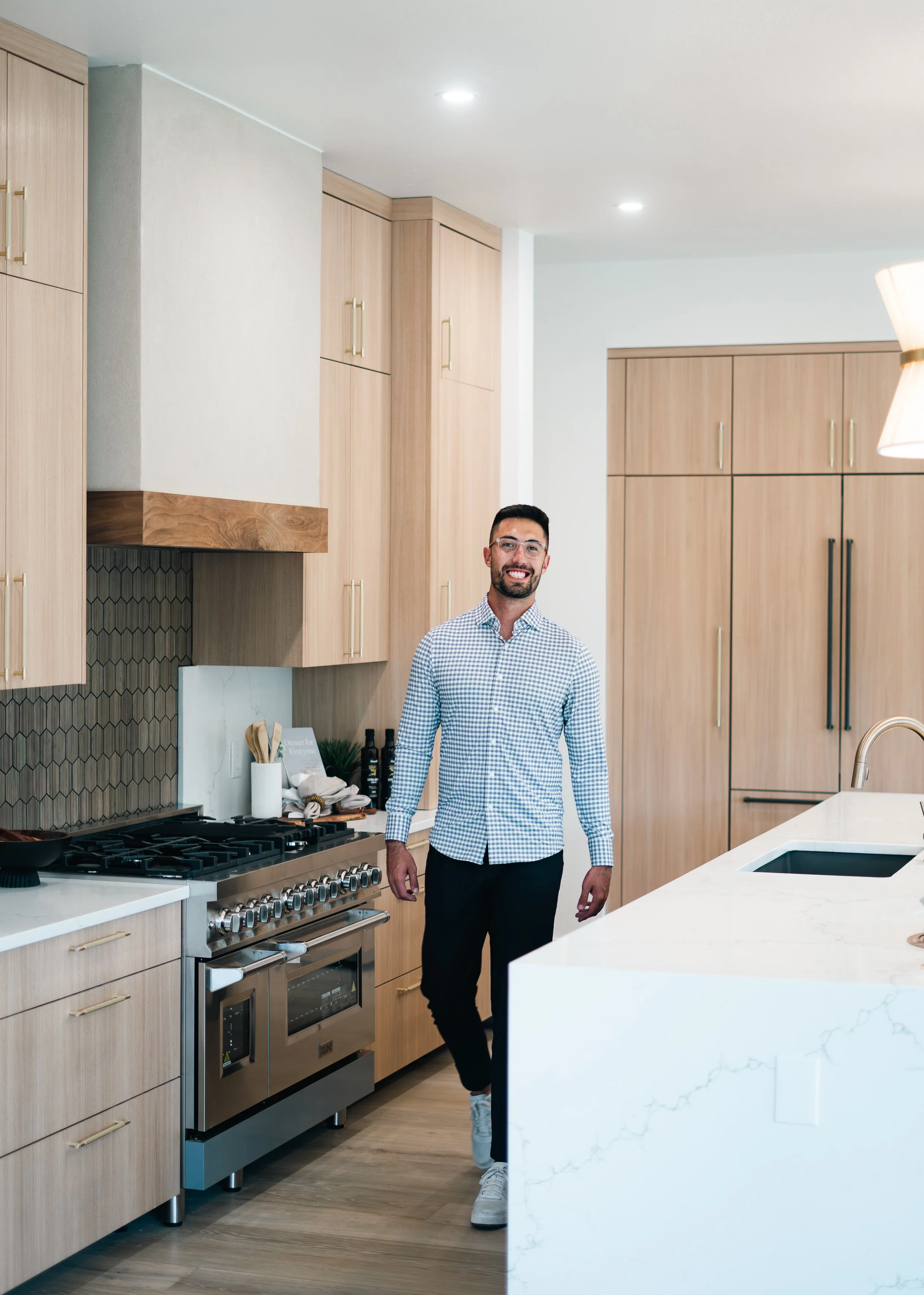 Smiling man in checkered shirt and black pants standing in a modern kitchen with light wood cabinets and a white marble island.