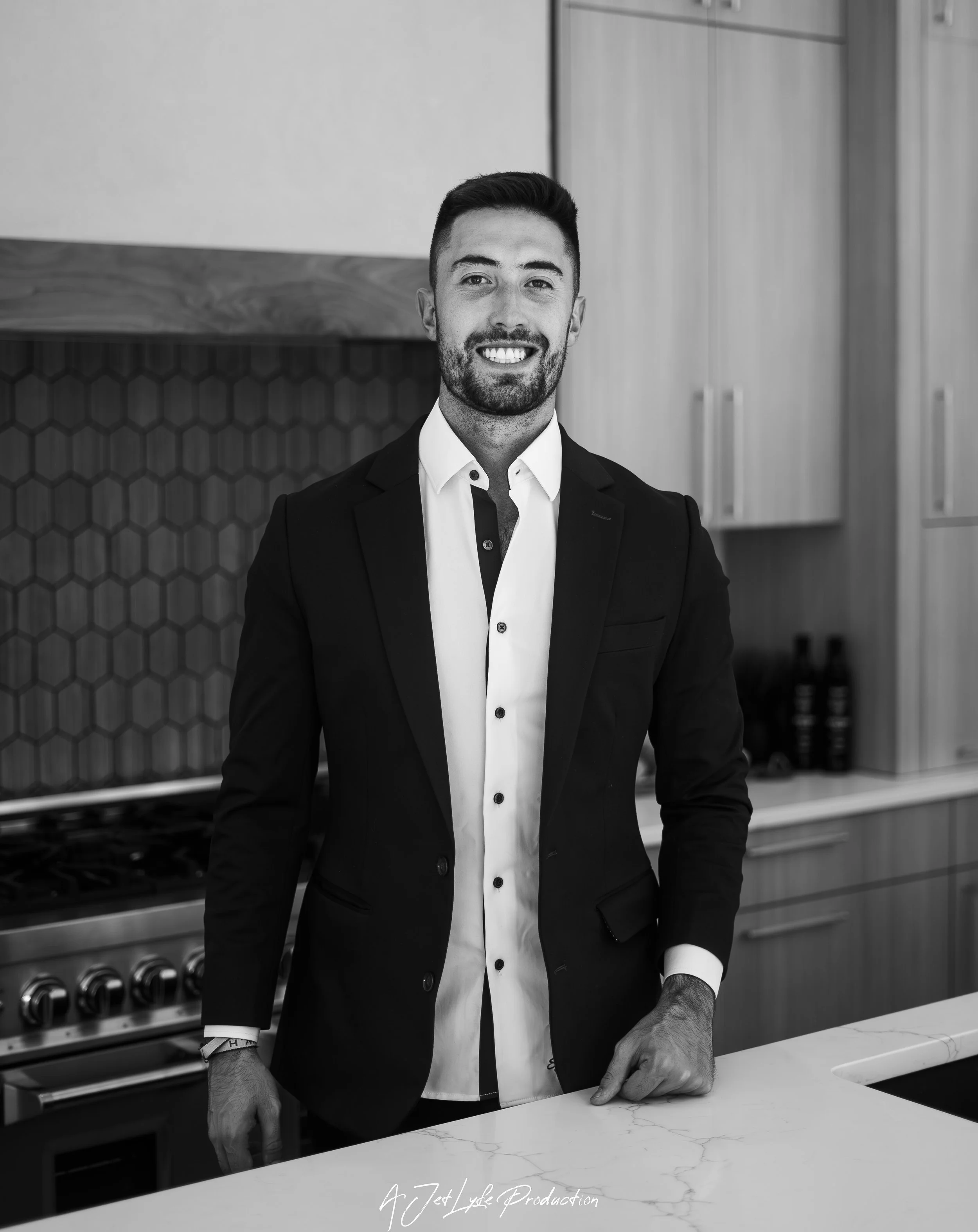 A smiling man in a suit standing in a modern kitchen with a stove and cabinets.