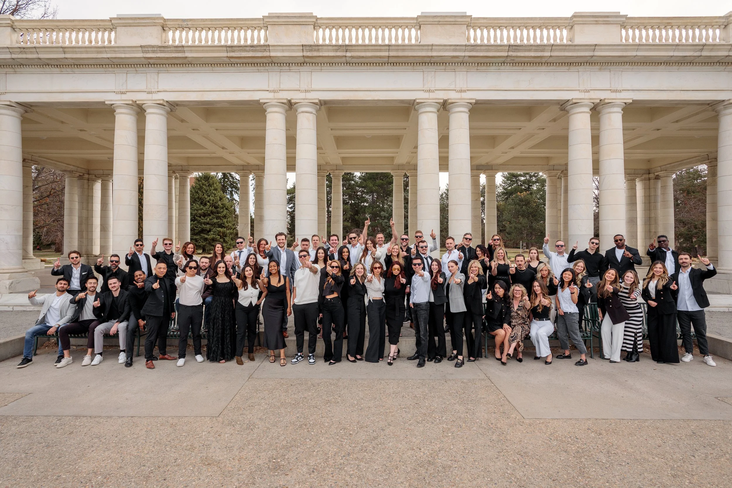 A large group of people dressed in formal and semi-formal attire, standing and sitting in front of a classical-style building with tall columns and balustrades.