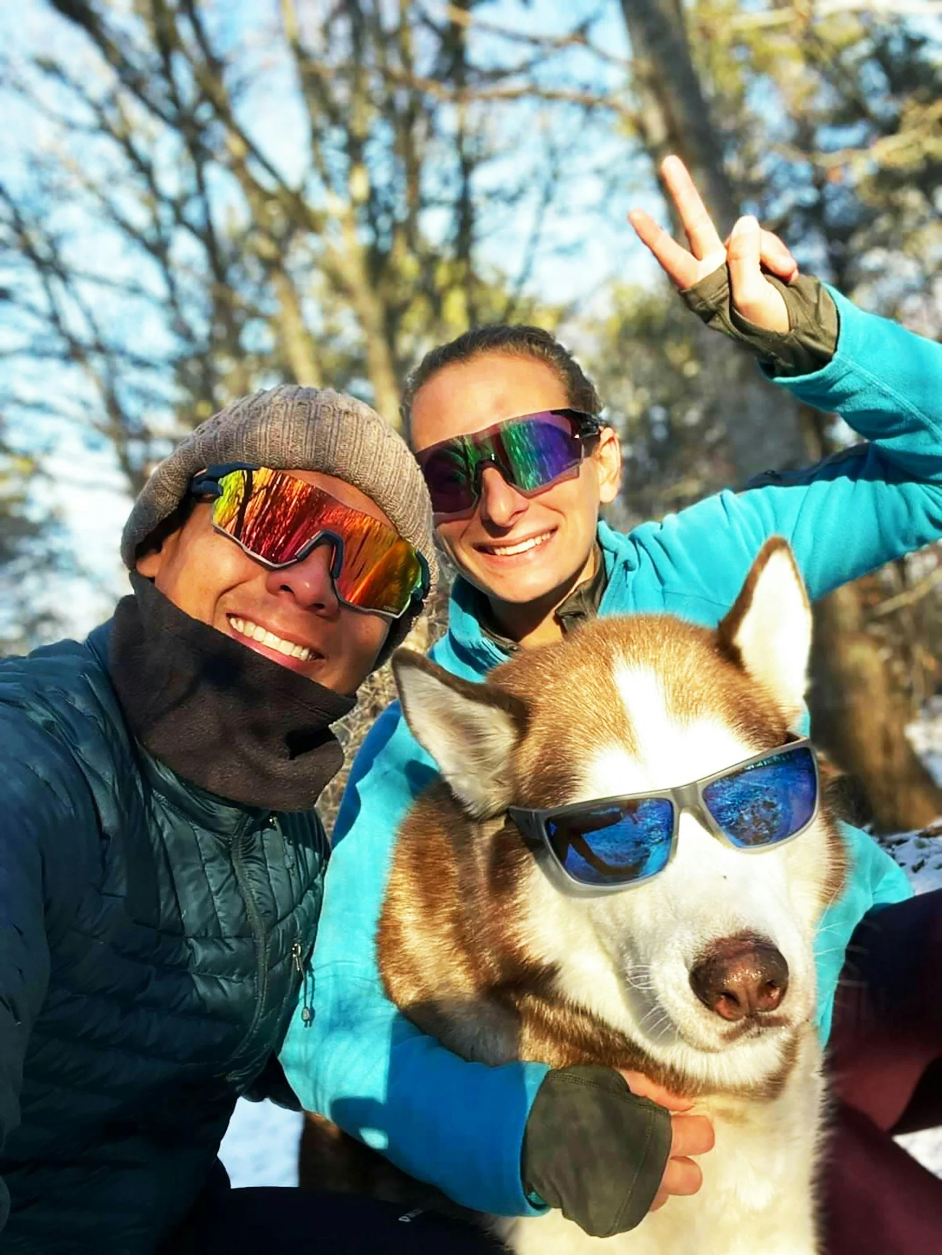 Two people wearing sunglasses and outdoor jackets, smiling, with a dog wearing sunglasses, in a snowy outdoor setting with trees in the background.