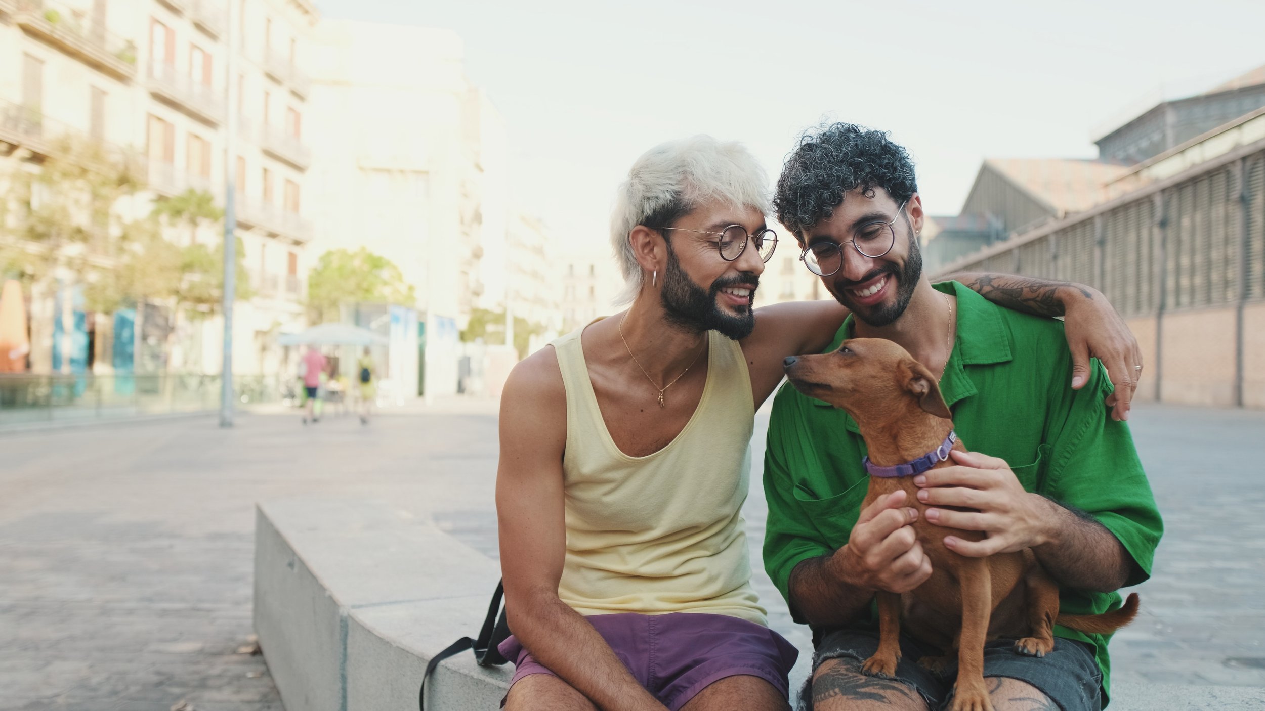 Two men with glasses and beards sitting on a bench in an urban area, smiling and interacting with a small brown dog with a purple collar.