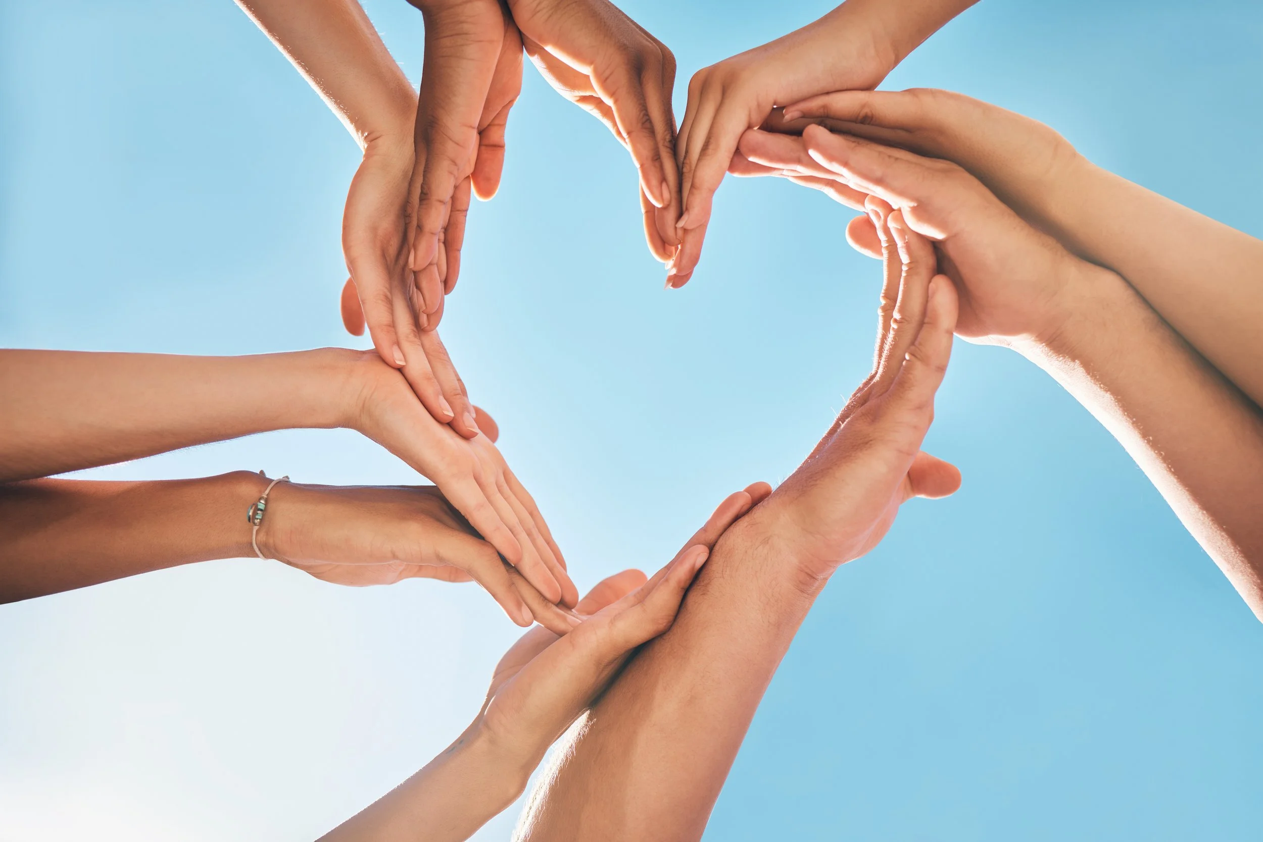 Multiple hands forming a heart shape against a blue sky.