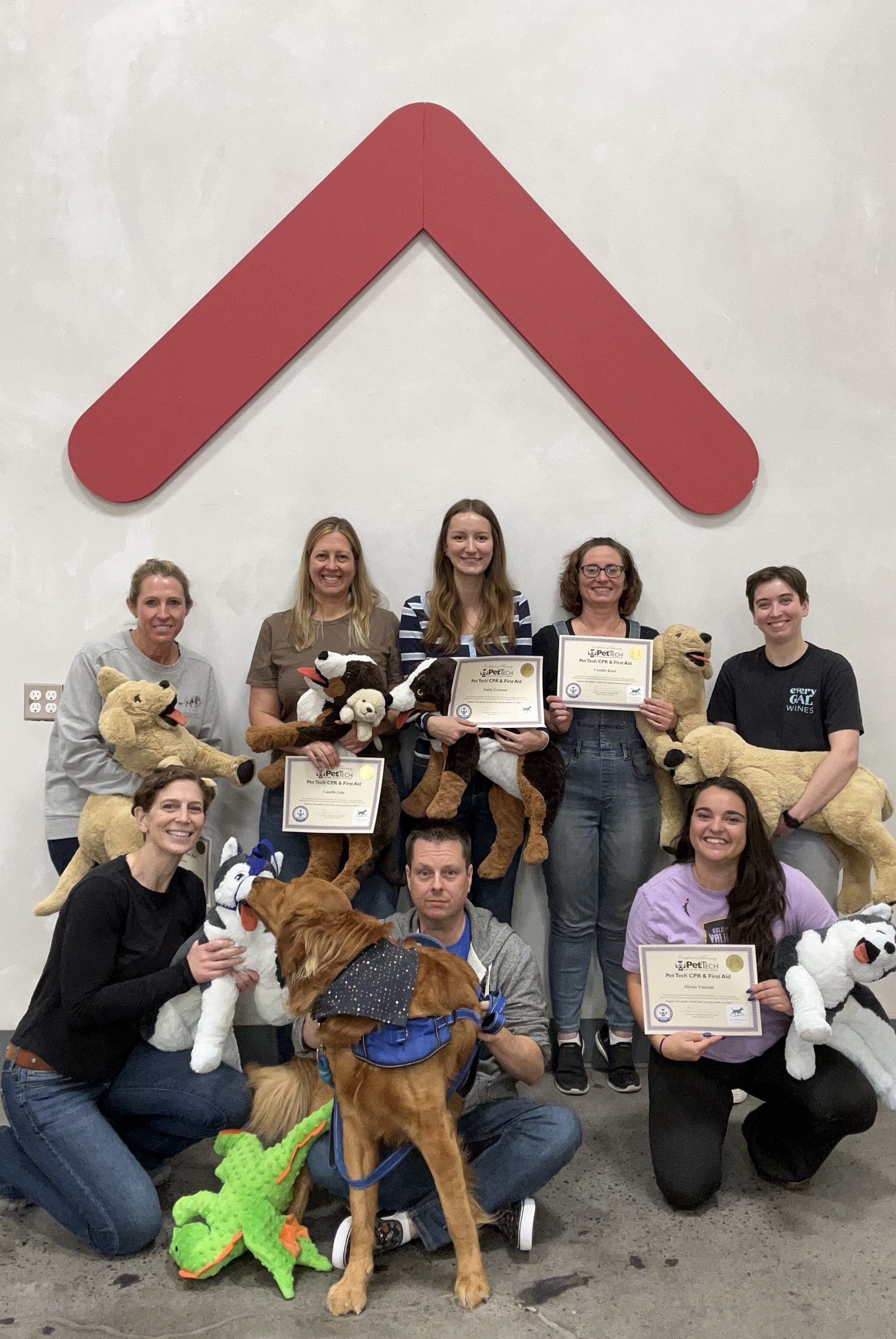A group of people holding certificates and stuffed animals, with some dogs in the foreground, standing in front of a large red arrow on the wall.