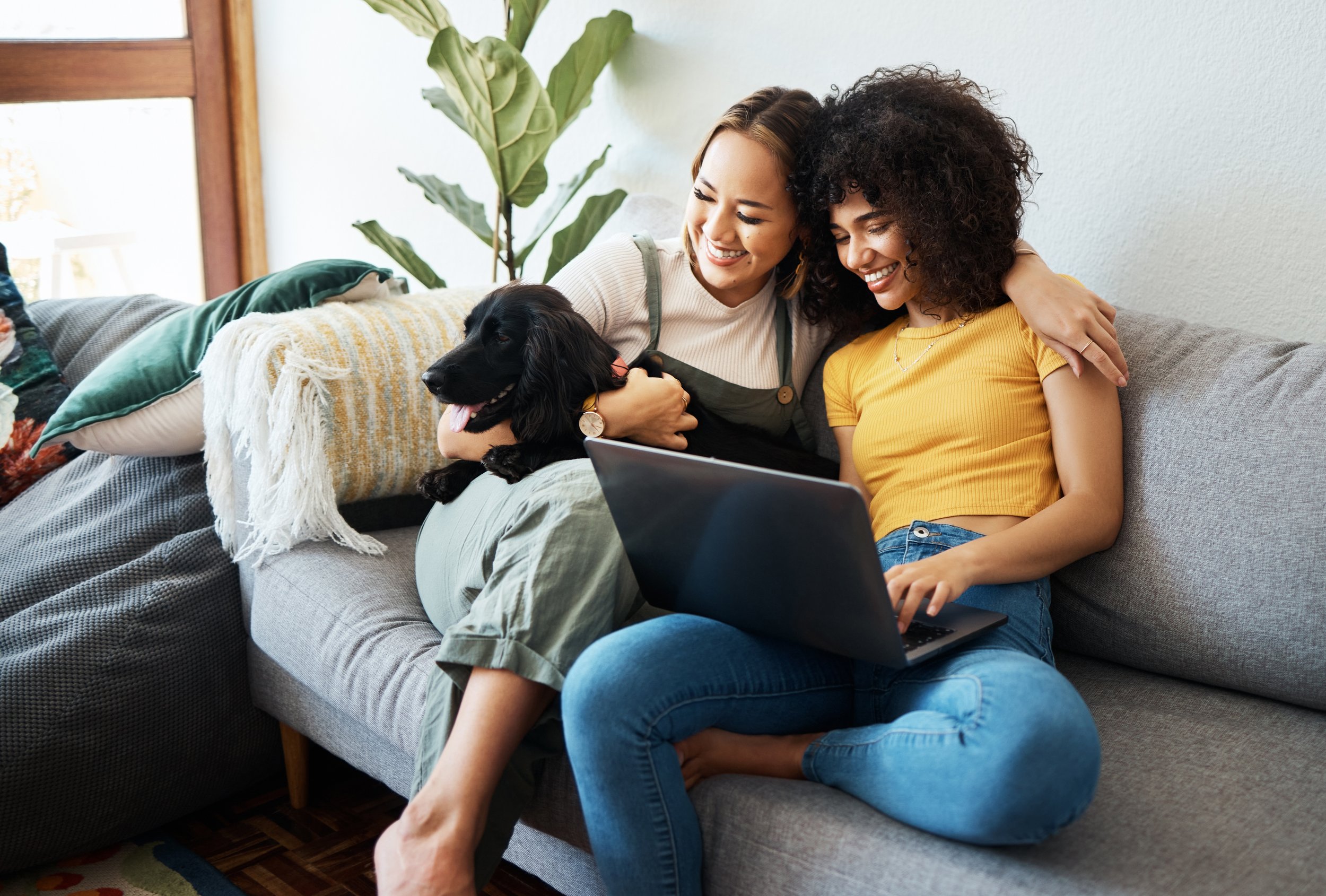 Two women sitting on a sofa, smiling and hugging each other, with a black dog on their laps, in a bright living room with a large plant in the background.
