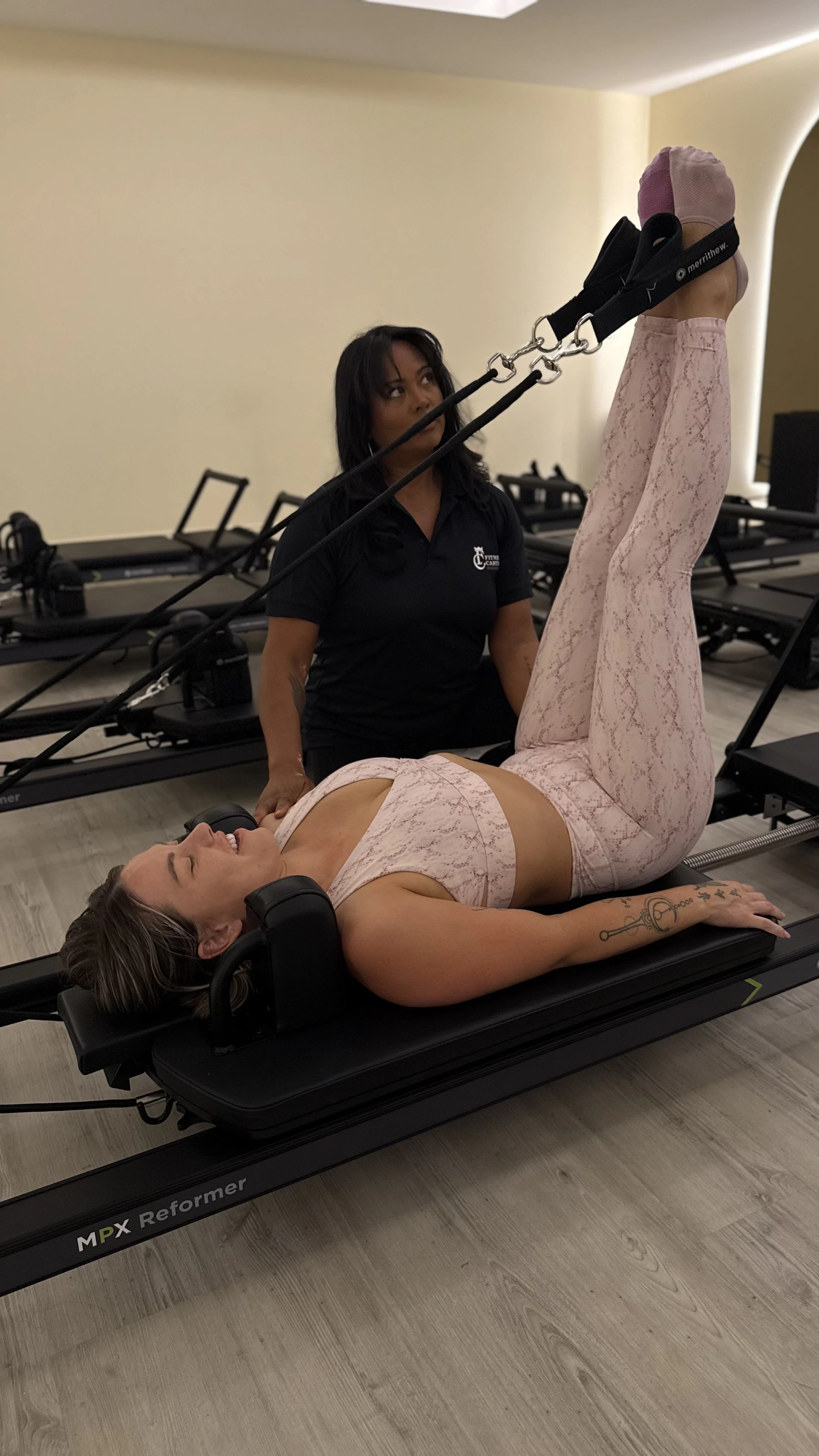 A woman lying on a Pilates reformer machine, smiling, with her legs extended upward and holding onto straps. A trainer stands nearby, observing her.