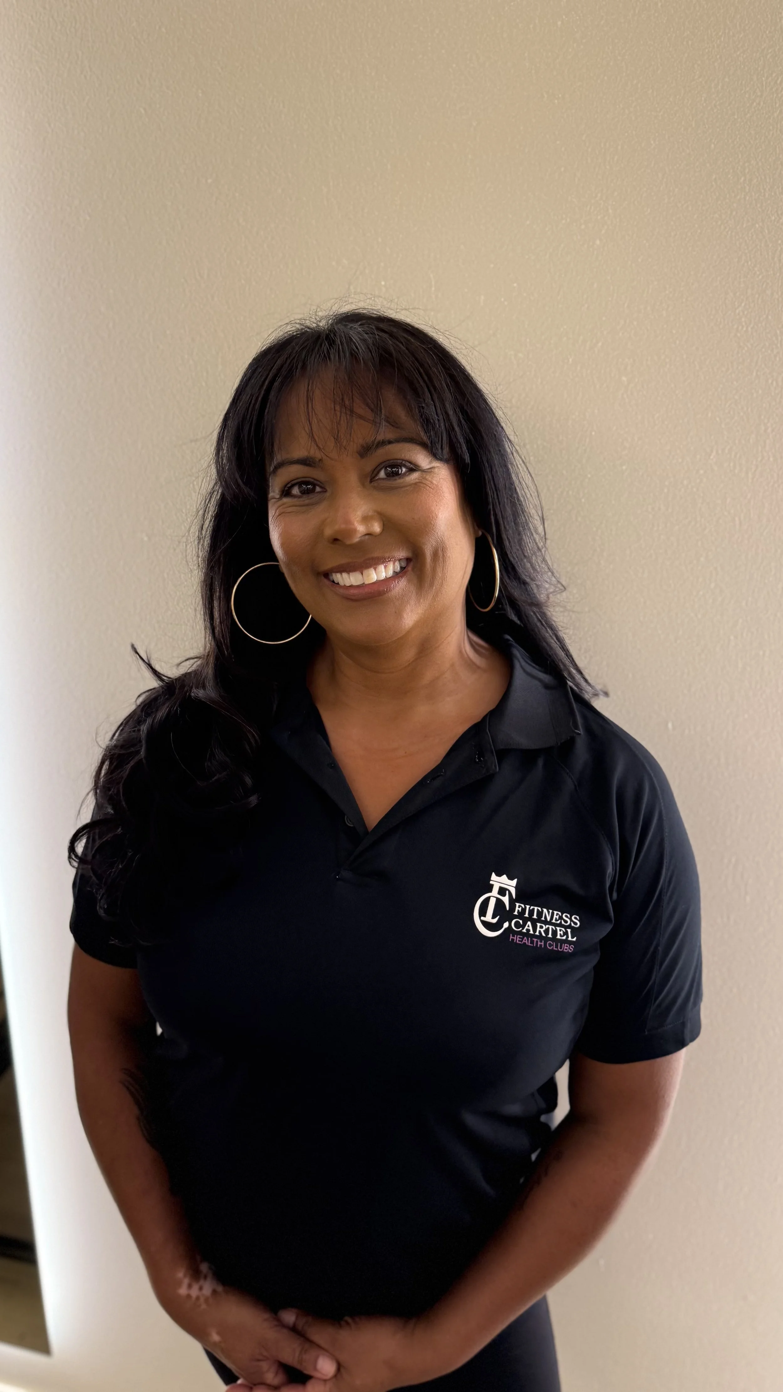 Smiling woman with hoop earrings wearing a black polo shirt with 'Fitness Cartel Health Clubs' logo, standing against a beige wall.