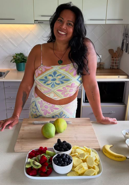 Smiling woman with dark hair in a kitchen, standing behind a counter with a wooden cutting board, two green apples, and a platter of fresh fruit including strawberries, kiwi, blueberries, blackberries, pineapple, and grapes, with a banana on the side.