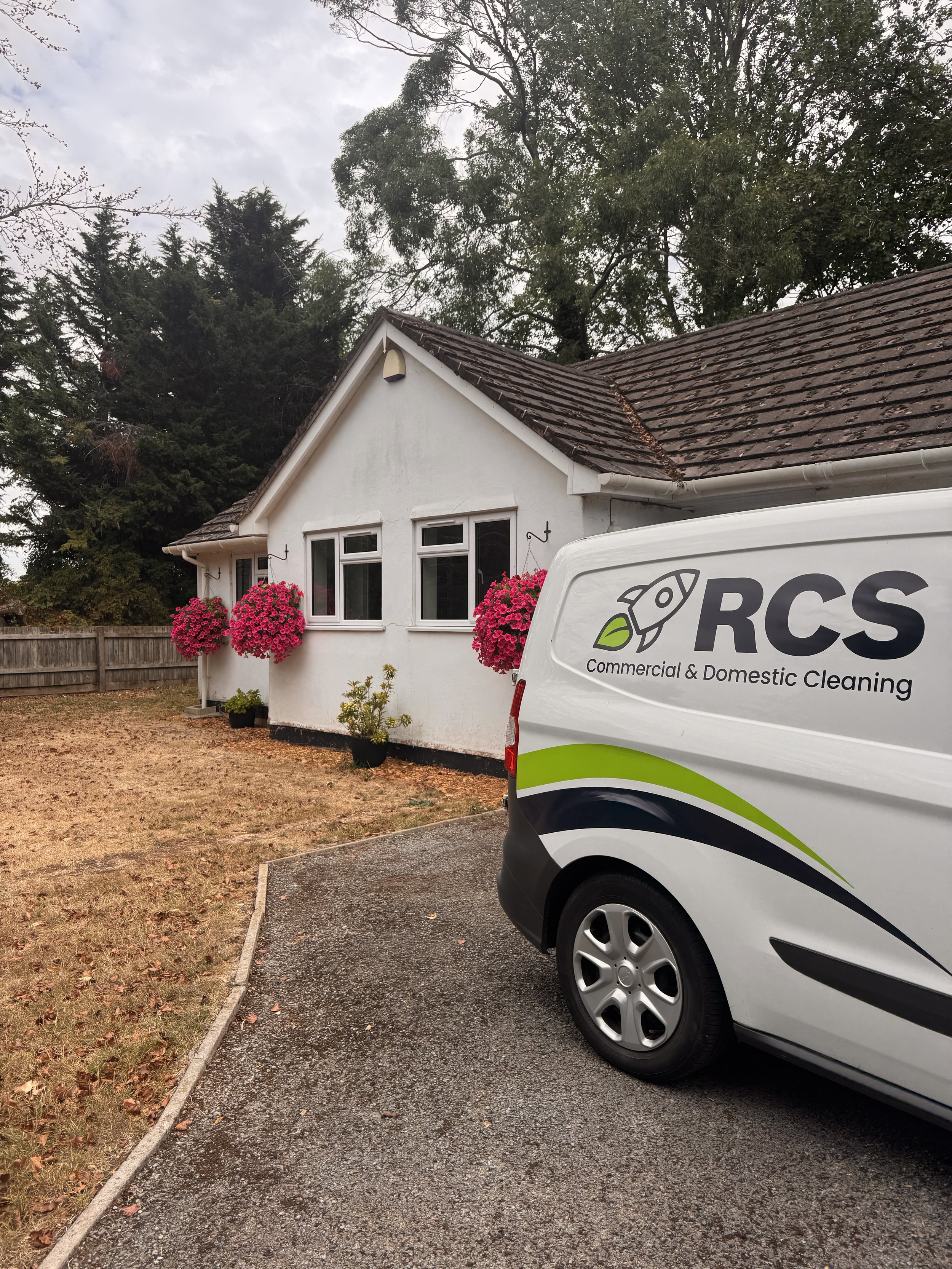 A white van with RCS Commercial & Domestic Cleaning logo parked in a driveway in front of a house with flower baskets.