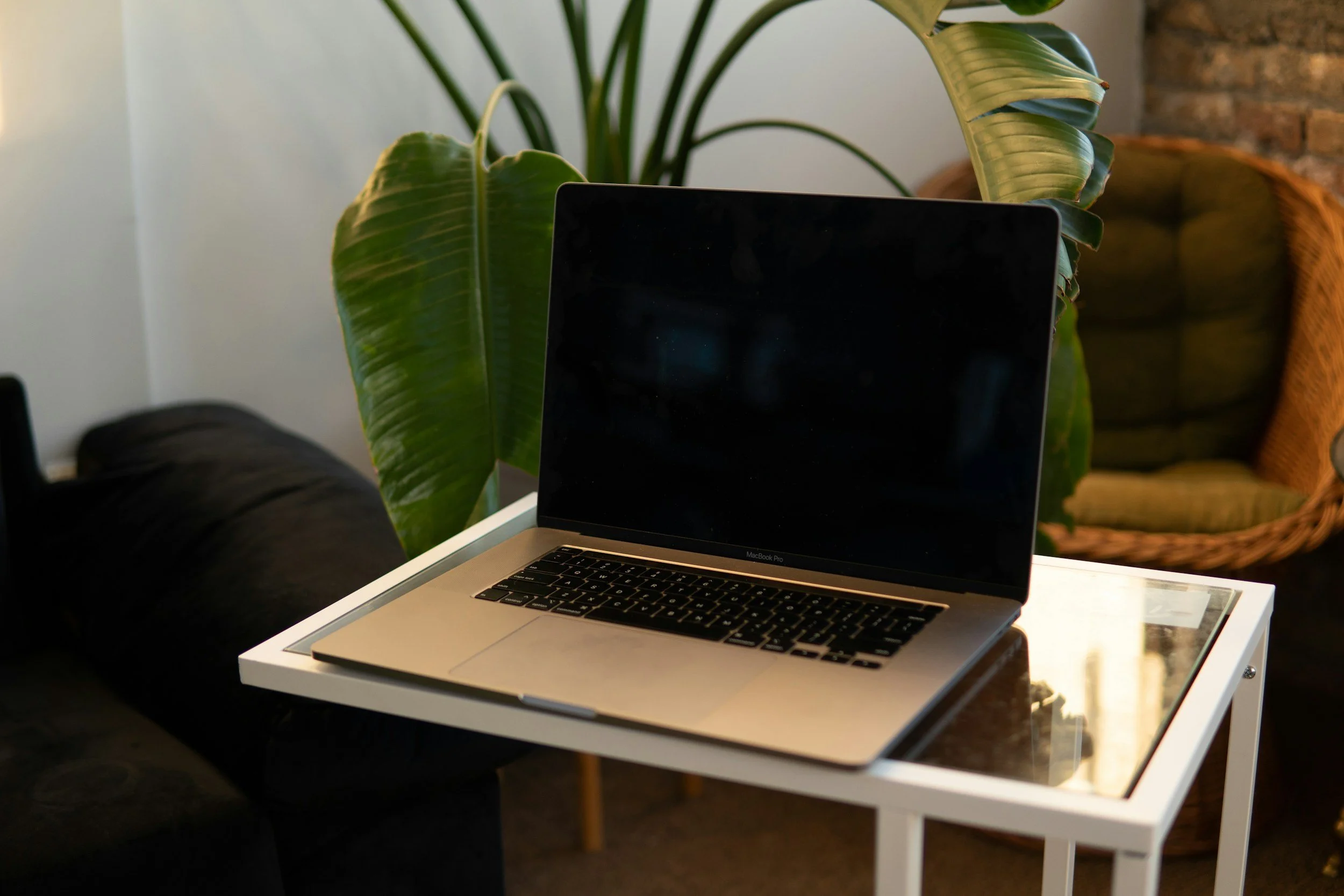A MacBook Pro on a white side table with a glass top, positioned in front of a black person sitting on a dark-colored couch. Behind the table, there are large green tropical plant leaves and a wicker chair with green cushions in a cozy, stylish room.