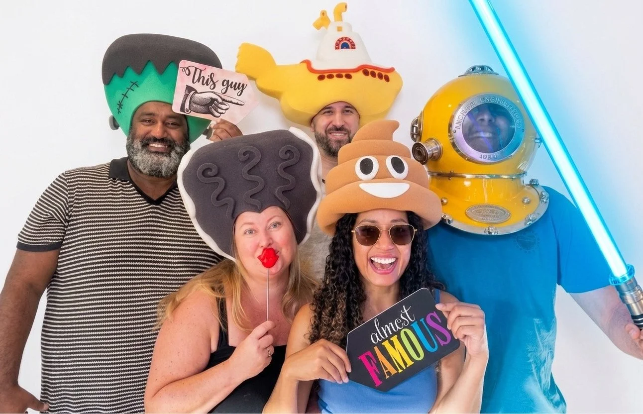 A group of six friends wearing humorous costumes and holding funny signs, posing in front of a pink sequined backdrop at a photo booth.