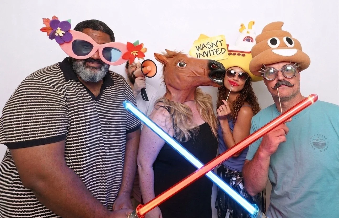 Group of six friends in funny costumes and holding humorous signs, standing in front of a pink sequin backdrop. One person wears a horse head mask, another a poop emoji hat, one a unicorn hat, and another star-shaped sunglasses. They are laughing and smiling.