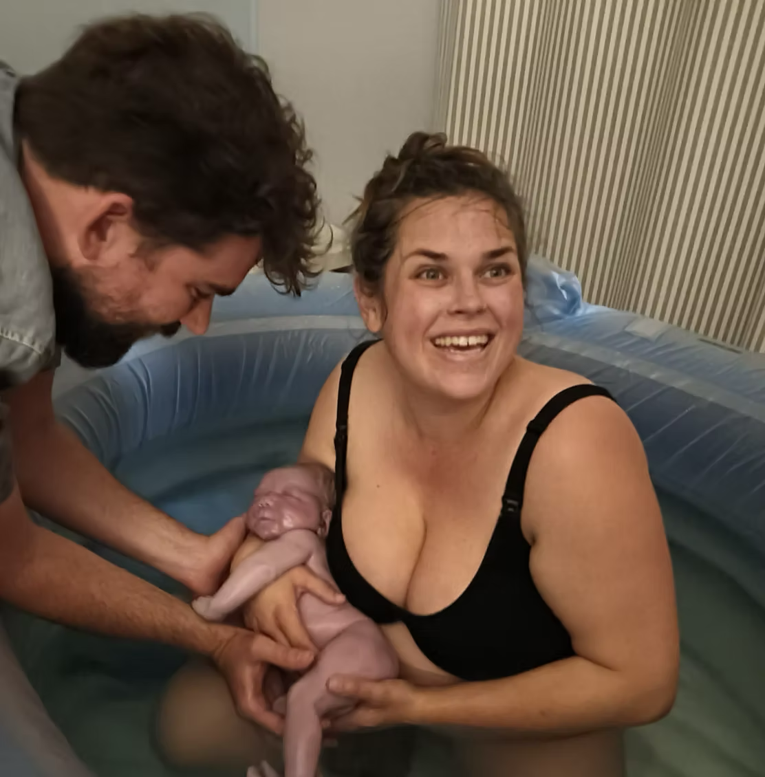 Picture of a person in a birthing pool holding a newborn in their hands. Their partner is outside the pool and is gently holding the baby too