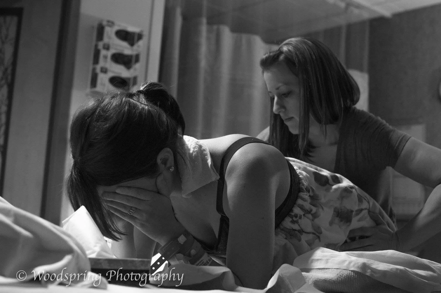 A woman in labor on a hospital bed, with another woman supporting her during childbirth, in a hospital room setting.