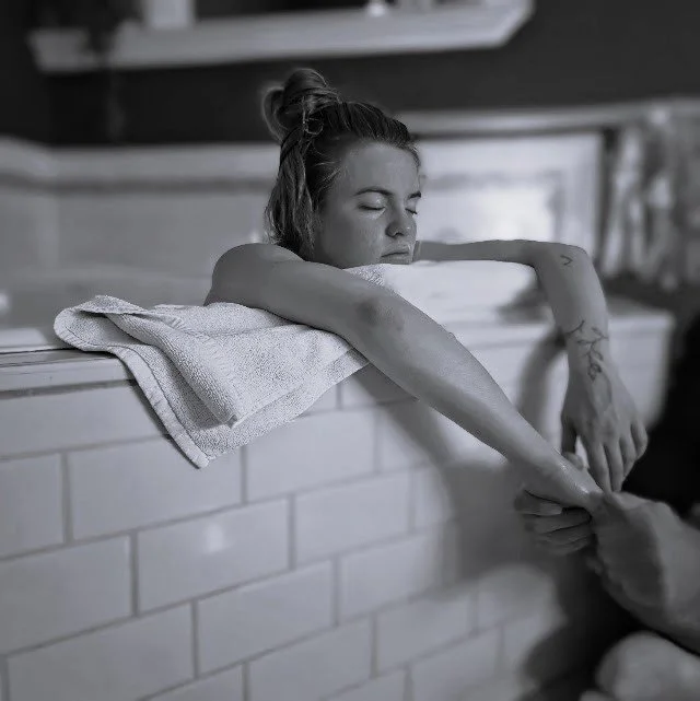 A woman enjoying a relaxing bath, resting her arms over a towel-draped edge of the bathtub with her eyes closed.