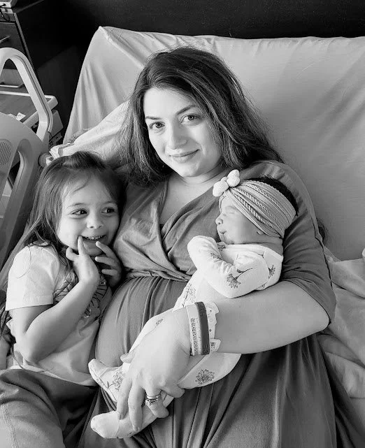 Woman in a hospital bed holding newborn baby with a young girl nearby, both children smiling.