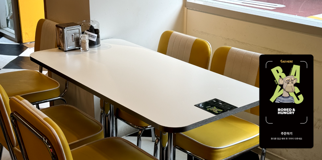Empty booth-style dining table with yellow chairs in a casual restaurant, near a window, with condiments and napkin holder on the table, and a promotional sign on the table that says "BORED & HUNGRY" with cartoon character and text in Korean.