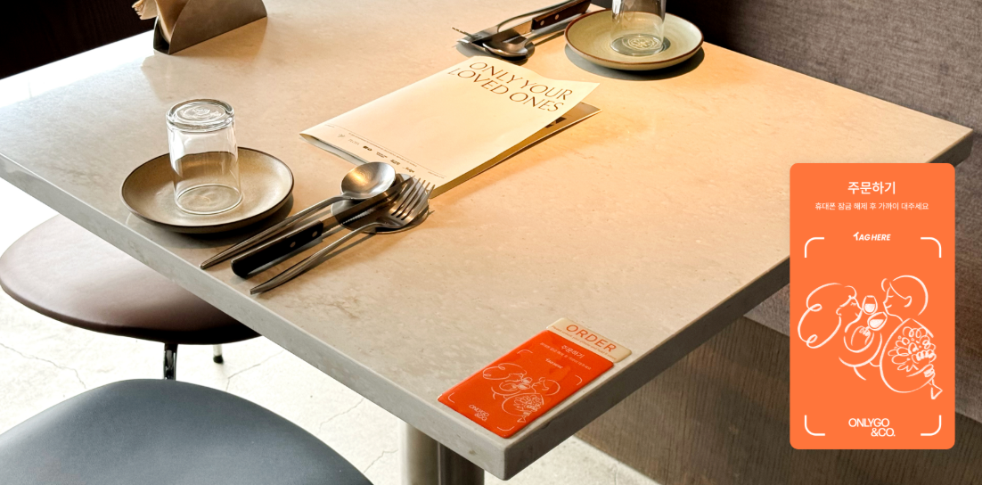 Empty restaurant table set with a glass, a plate, a spoon, a fork, and a knife. There is a paper menu, a small orange order card, and a side of snacks illustration on an orange card on the table.