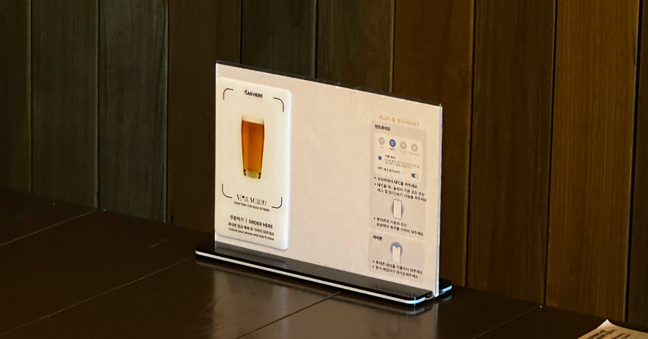 Table sign with a picture of a beer glass and text instructing to order here, placed on a dark tabletop against a wood-paneled wall.