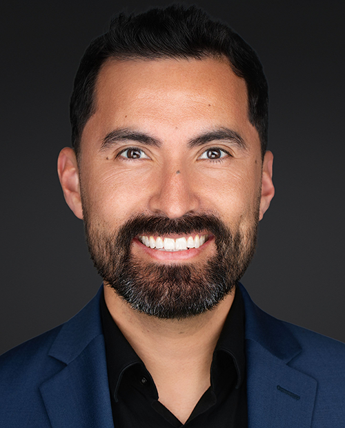 Headshot of a smiling man with dark hair, beard, and mustache, wearing a dark blue suit jacket and black shirt against a dark gray background.