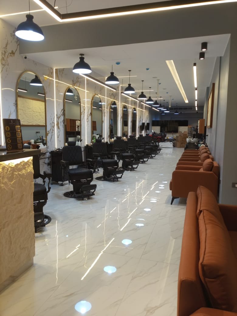 A modern salon interior with black barber chairs aligned against a marble wall with gold accents and large arched mirrors, plush brown seating along the right wall, marble flooring, and black pendant lights hanging from the ceiling.
