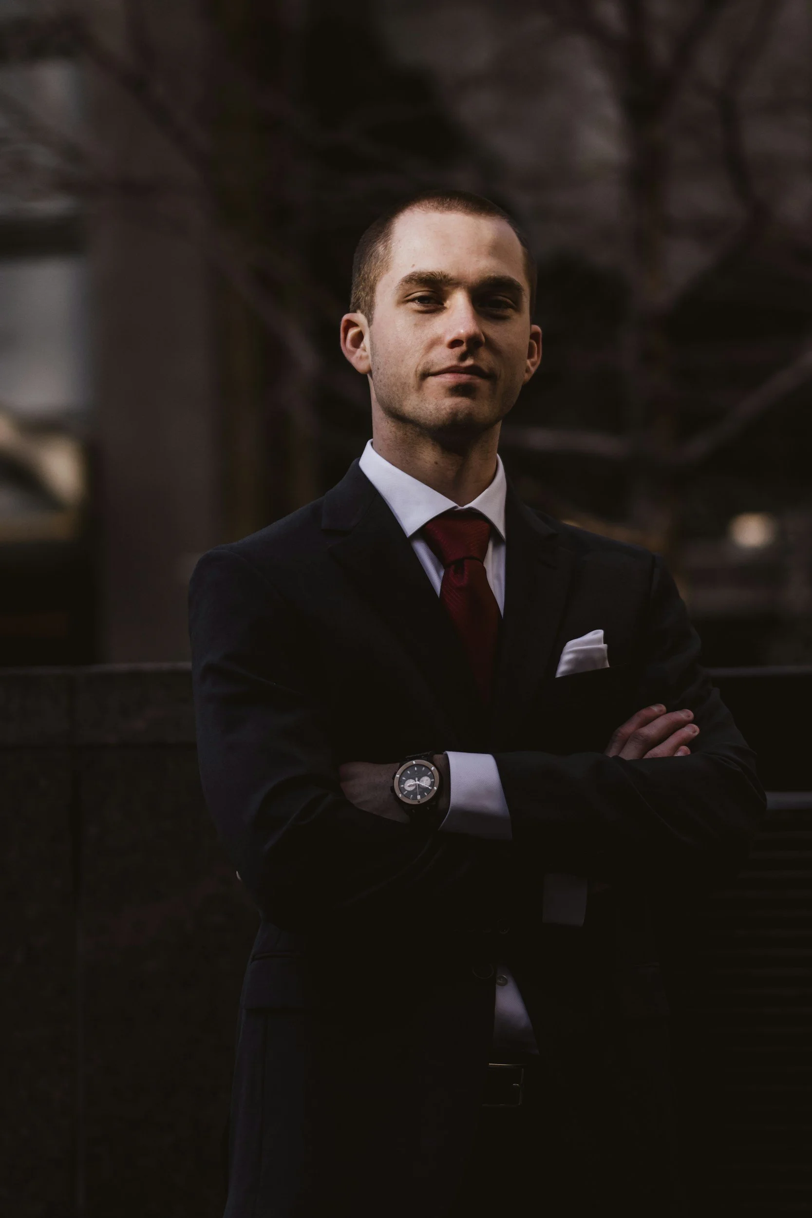 A young man in a dark suit with a white shirt and red tie, standing outdoors with arms crossed, wearing a watch on his left wrist, with blurred trees and building in the background.