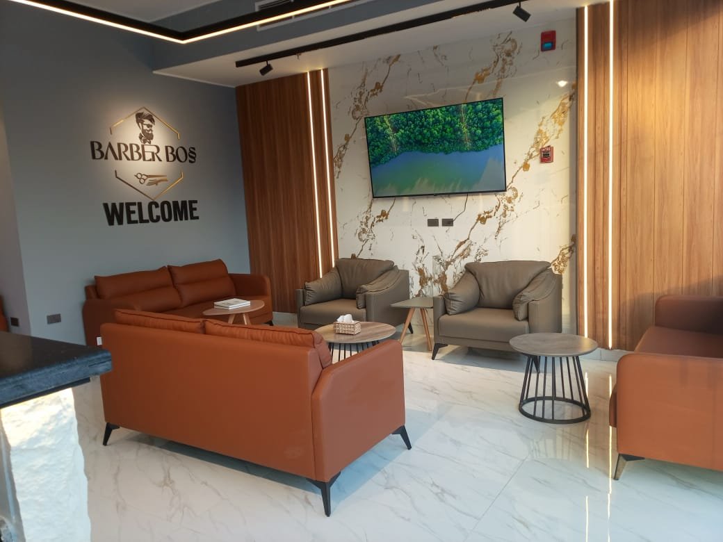 Modern lounge area with gray and brown armchairs, a leather sofa, a wall-mounted TV, a reception desk, and decorative wooden panels with vertical lighting. There is a welcome sign on the wall reading "Barber Bos Welcome".