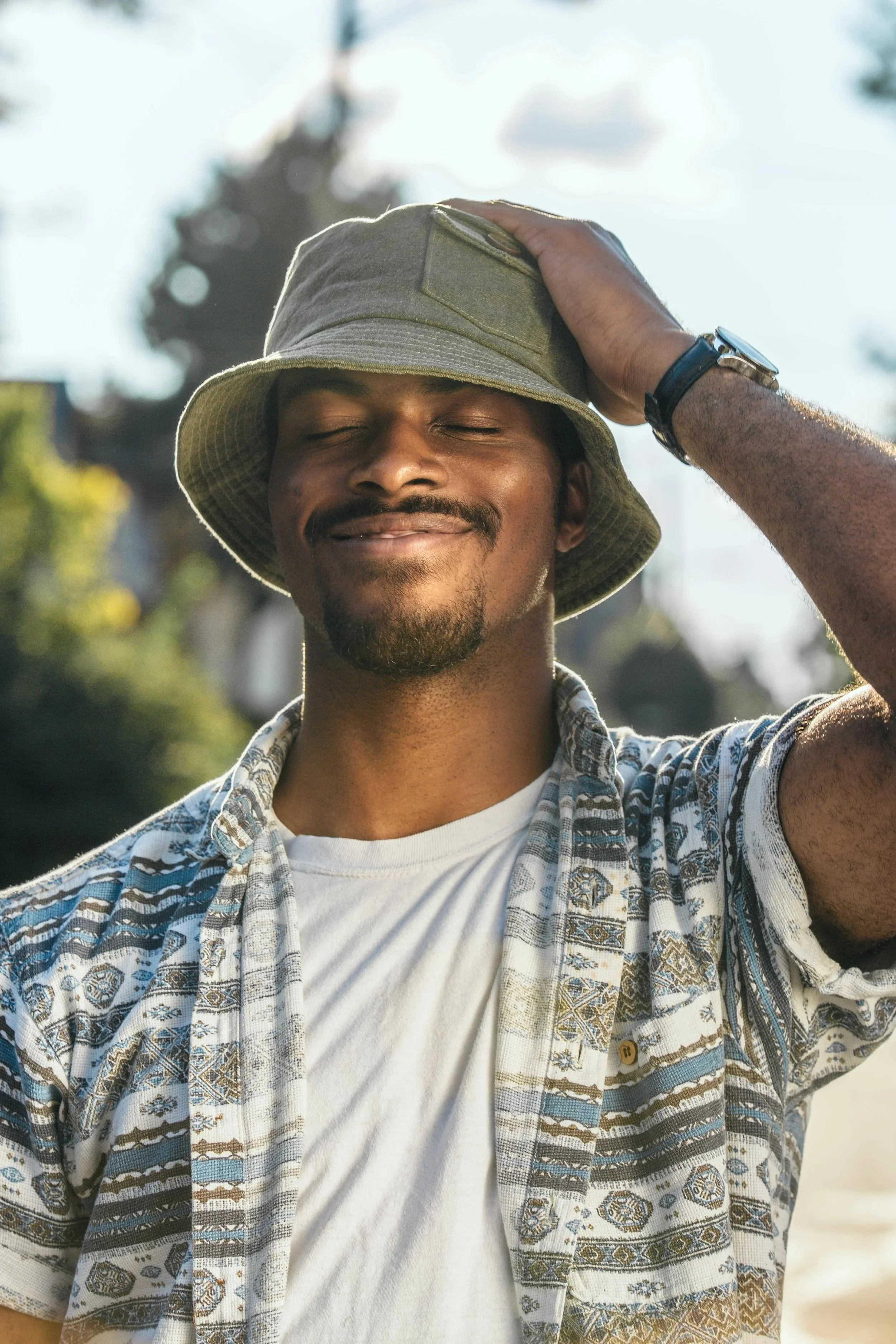 A man smiling with eyes closed, wearing a beige hat, a patterned short-sleeve button-up shirt, and a white t-shirt, standing outdoors in sunlight.