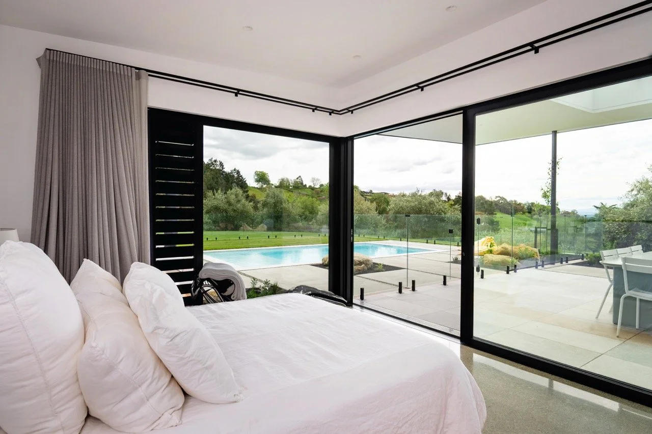 View of a bedroom with large glass sliding doors leading to a backyard with a swimming pool and green landscape.