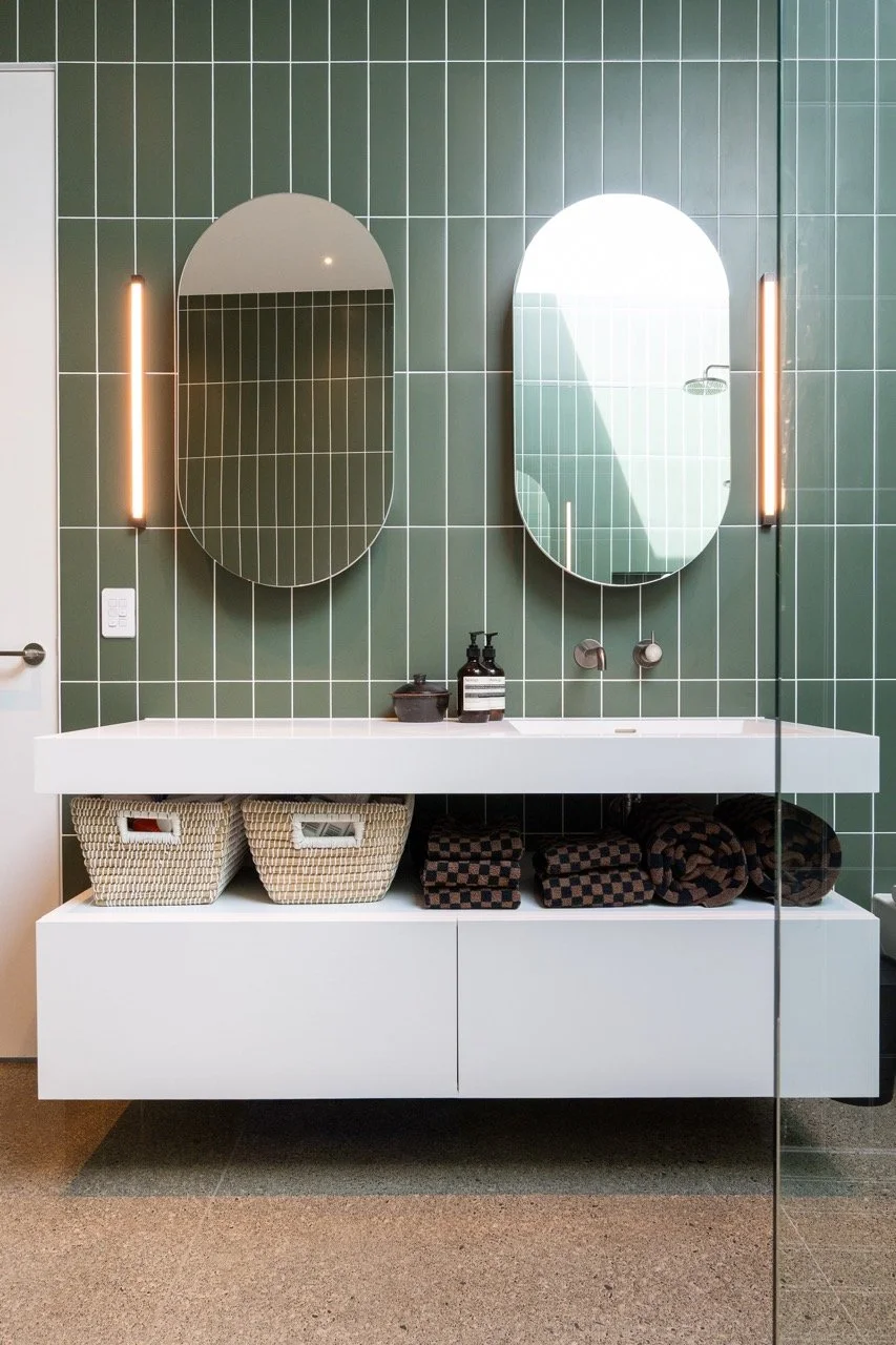 A modern bathroom vanity with two oval mirrors on green tiled wall, featuring a white floating cabinet with two woven baskets and folded towels underneath, black soap dispensers, and vertical light fixtures.