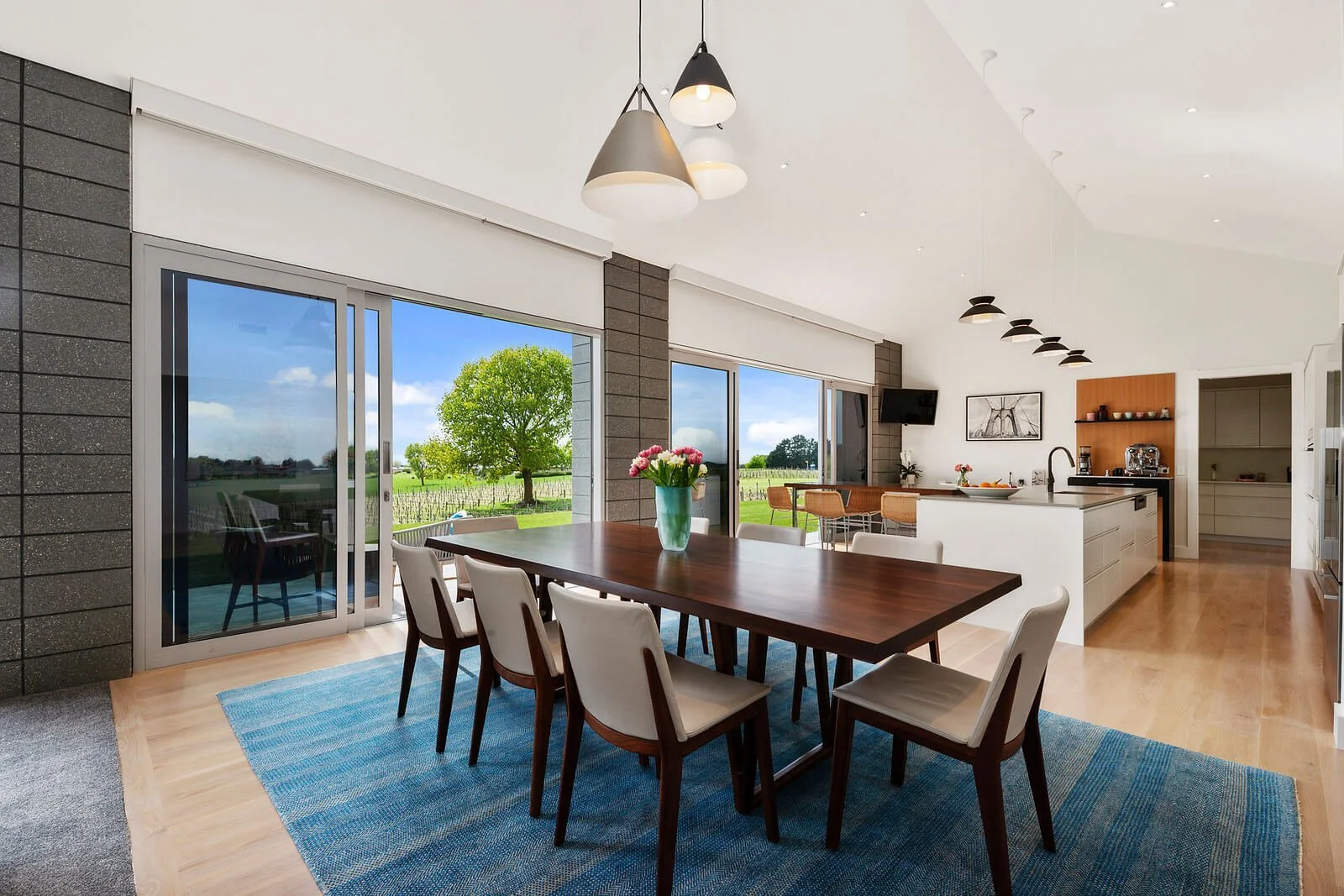 Modern kitchen and dining area with a large wooden table, white chairs, a light blue rug, and large glass sliding doors leading to an outdoor landscape with trees and a vineyard.