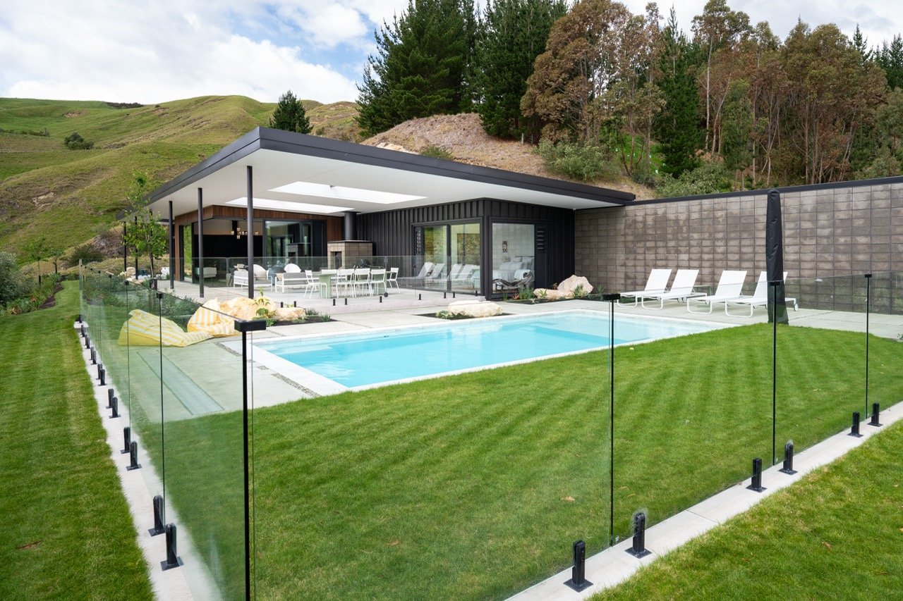 Modern house with a backyard pool, surrounding green grass, glass fence, and outdoor seating area, set against a hillside with trees.