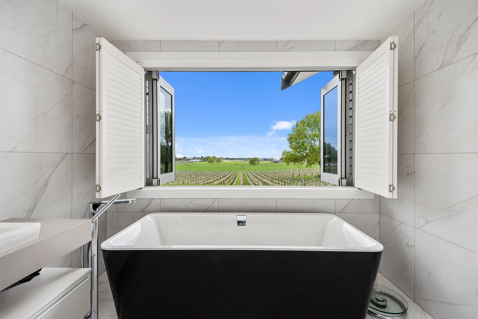 Modern bathroom featuring a black bathtub positioned in front of an open window with white shutters, revealing a scenic view of a green landscape with fields, trees, and a clear blue sky.