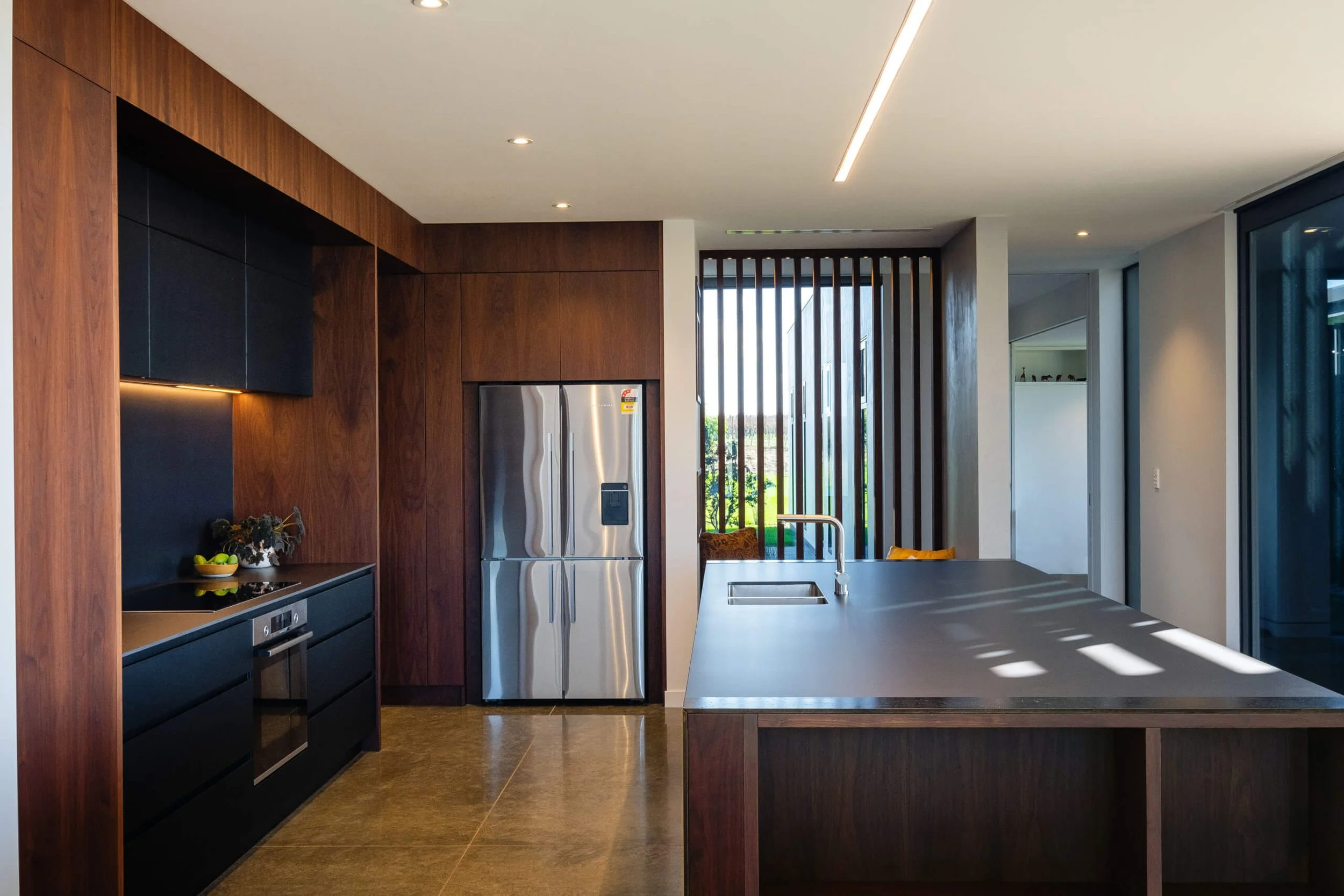Modern kitchen with wooden cabinets, stainless steel refrigerator, black appliances, and a large island with a sink, sunlight streaming through window slats.