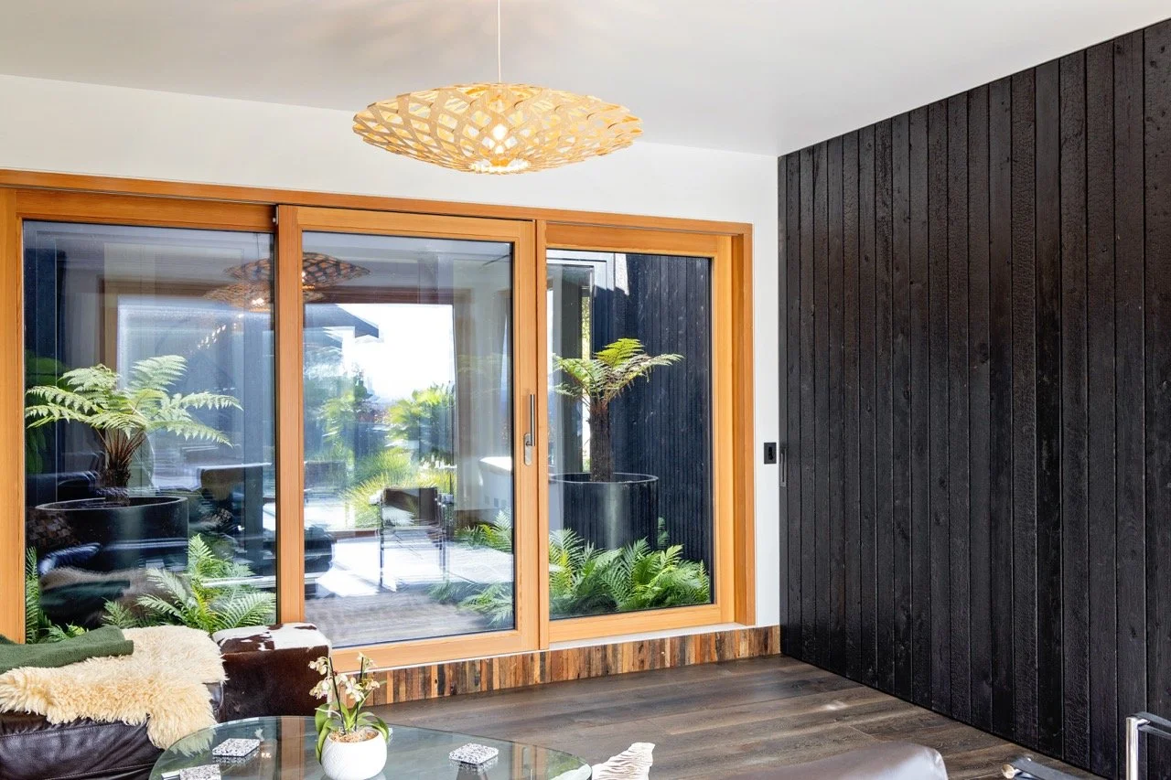 Living room with black wooden accent wall, glass sliding door with wooden frame, potted plants outside, modern beige pendant light, and a cozy seating area with a glass coffee table and cushions.