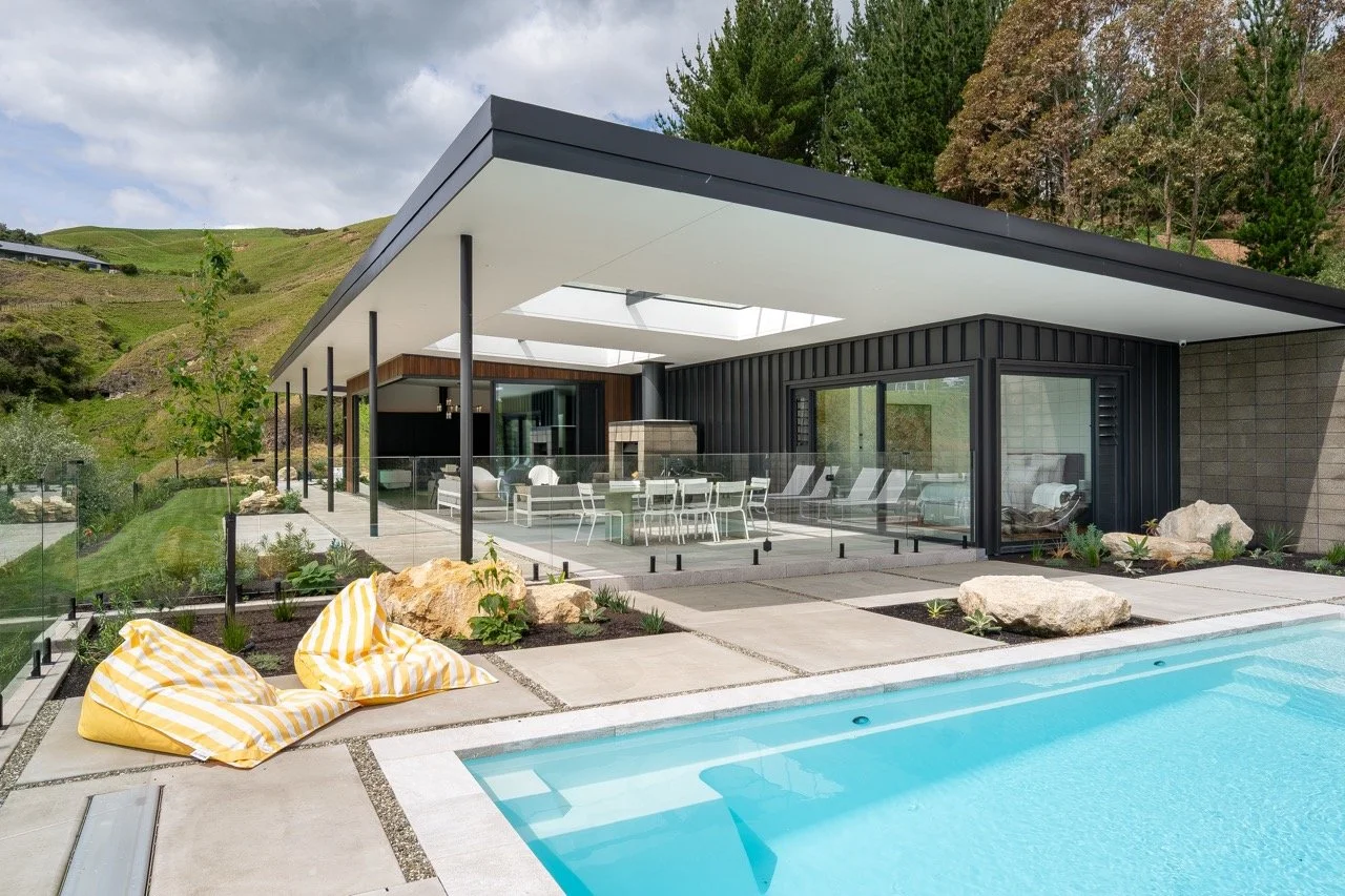 Modern house with outdoor pool, glass railing, and patio furniture, surrounded by landscaped garden and hillside.
