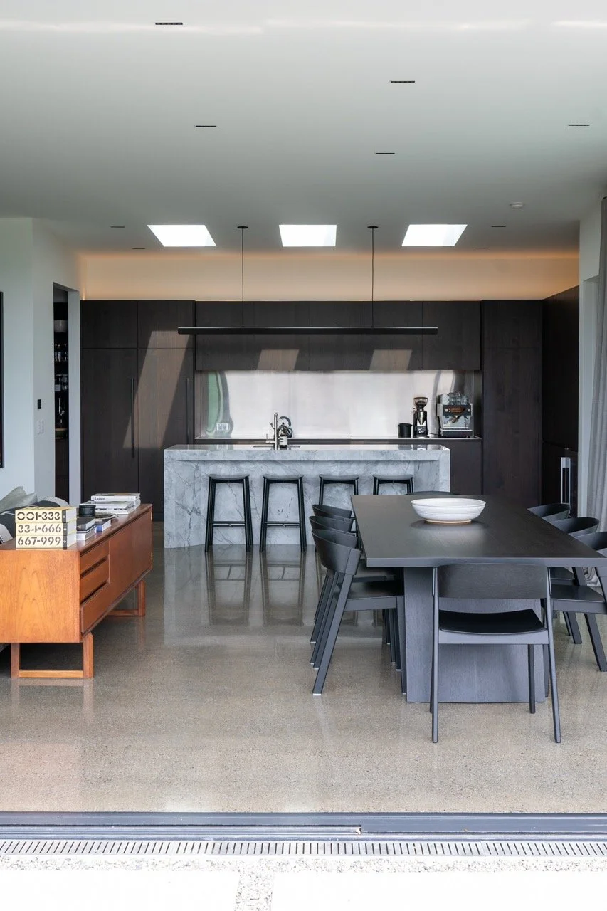 Modern kitchen with dark cabinets, white marble island, black dining table with chairs, coffee machine, coffee grinder, and a minimalist design.