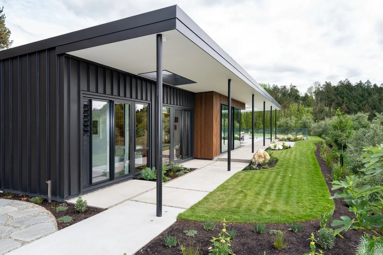 Modern house with black exterior, glass sliding doors, a covered patio supported by black poles, and a well-maintained green lawn with landscaped plants and trees in the background.