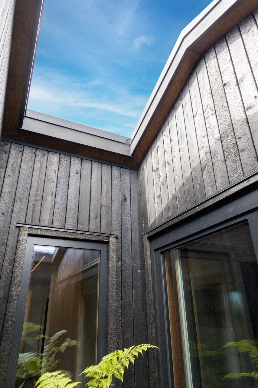 Corner of a building with black wooden siding, large glass windows, and an open skylight showing a blue sky with some clouds.