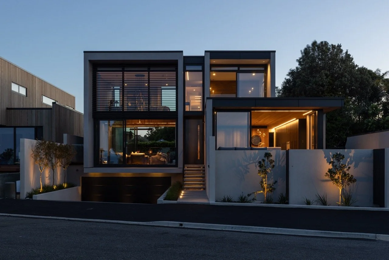 Modern two-story house with large glass windows, illuminated interior, and front yard with small trees and lighting