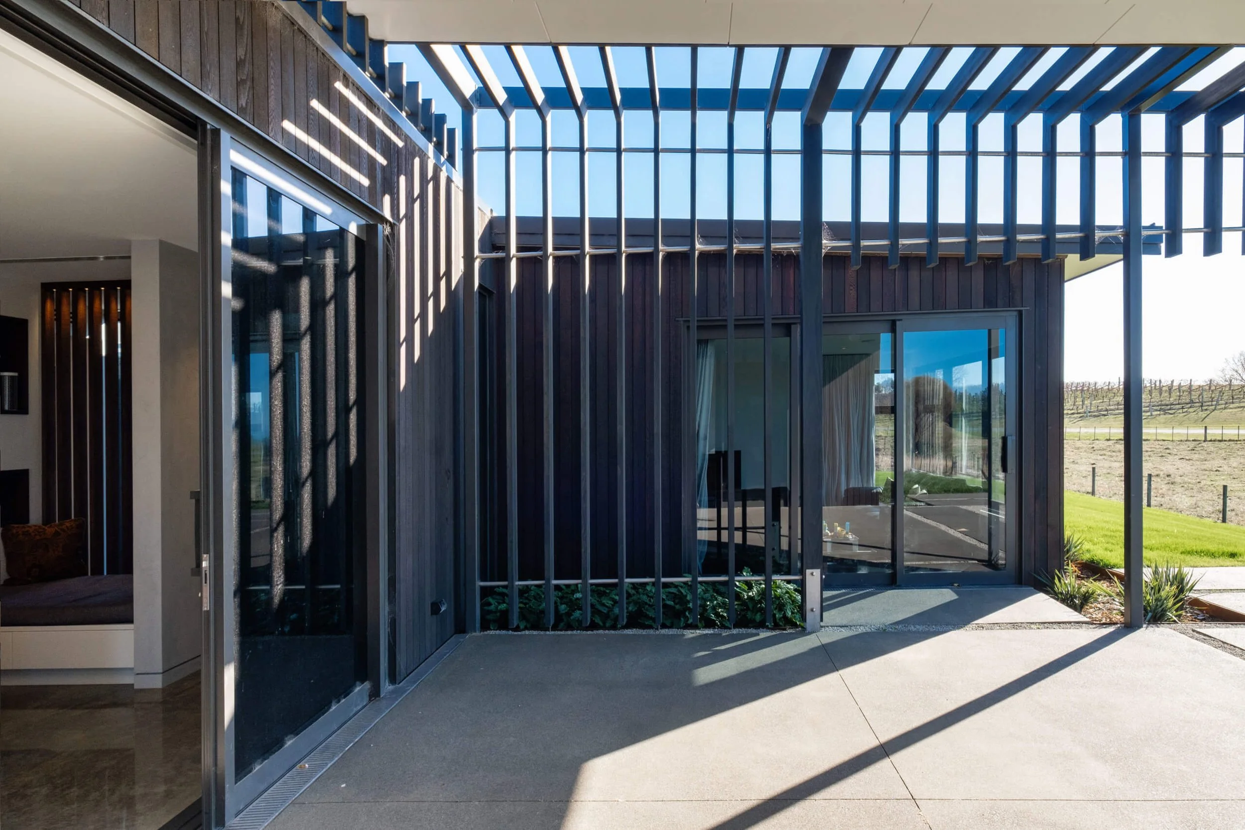 Exterior view of a modern house with wooden siding and large glass sliding doors, partially shaded by a pergola with slatted beams, overlooking a grassy area and distant vineyard.