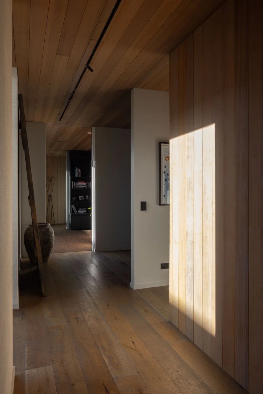 Interior hallway with wooden walls and ceiling, natural light casting a shadow on the wall, and a large decorative vase in the background.