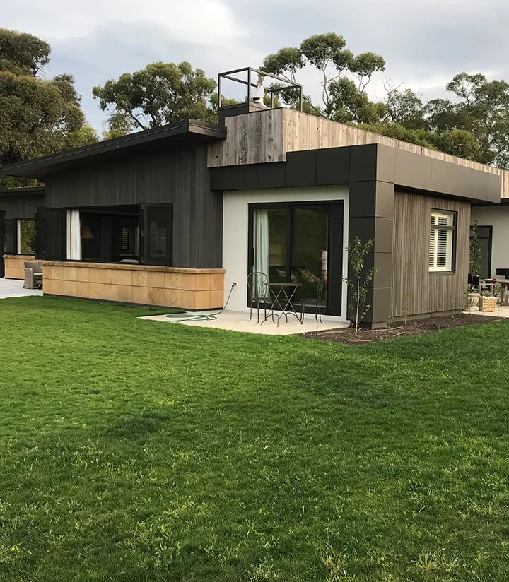 Modern house with dark and light gray exterior panels, large sliding glass doors, and a small patio with outdoor furniture, surrounded by a well-maintained green lawn and trees in the background.