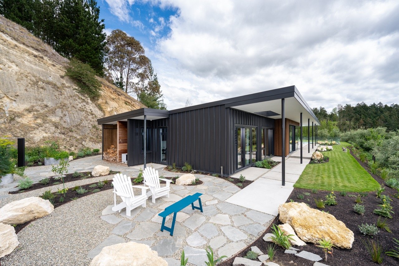 Modern house with black exterior siding, large glass doors, a covered porch with pillars, and a landscaped yard with rocks, plants, small trees, two white Adirondack chairs, a blue bench, and a gravel pathway, set against a hillside and cloudy sky.