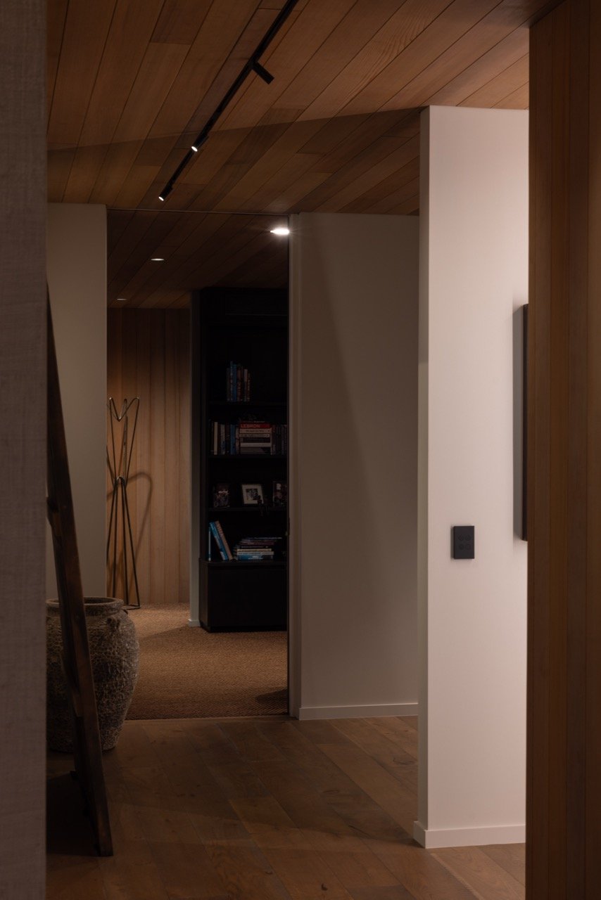 Interior view of a home with wood-paneled ceiling and floor, a white wall, and a dark bookshelf filled with books and framed photos.