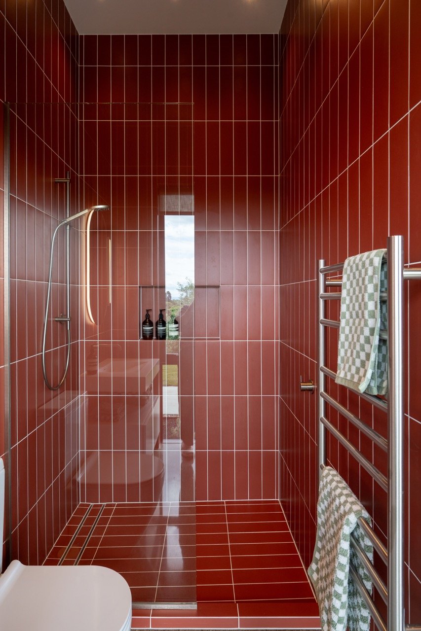 A modern bathroom with red tiles on the walls and floor, a walk-in shower with a glass door, a wall-mounted hand shower, a toilet, and a towel rack with two checkered towels.