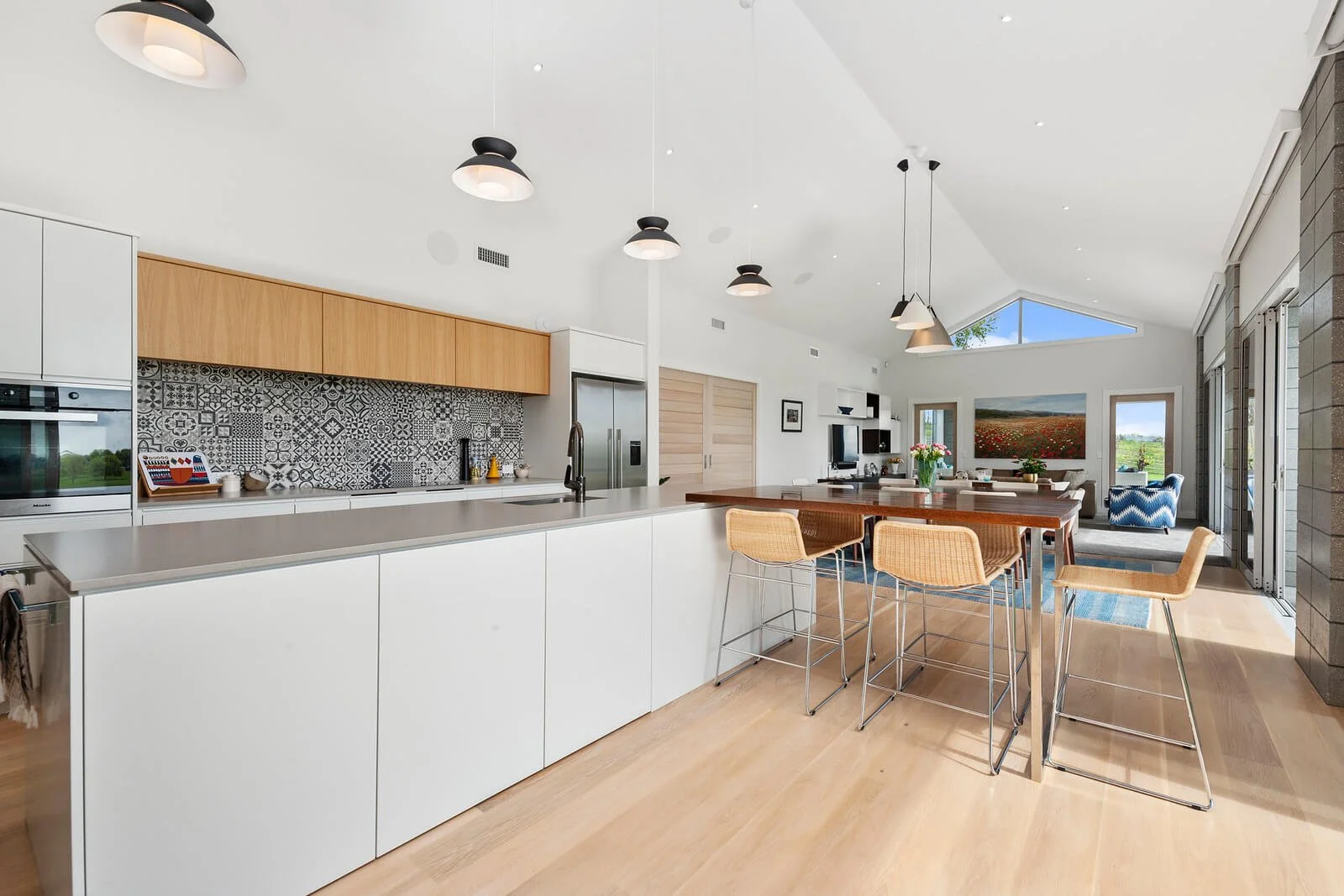 Open-concept kitchen and living room with modern design, white cabinetry, patterned backsplash, wooden accents, and a dining area with chairs around a table, large windows, and outdoor views.