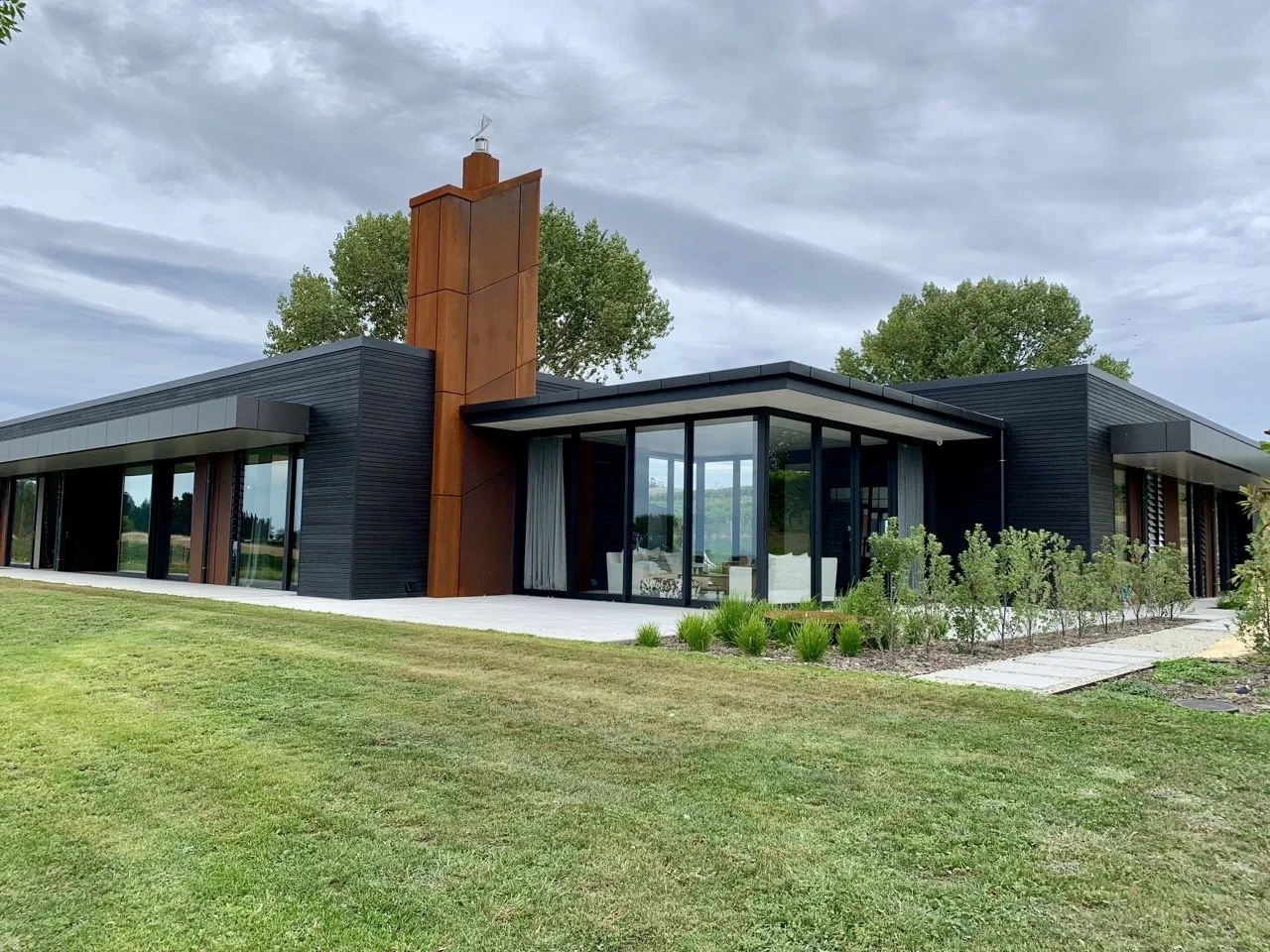 Modern single-story house with large glass windows, a black exterior, and a rust-colored chimney, surrounded by a well-maintained lawn and trees under a cloudy sky.