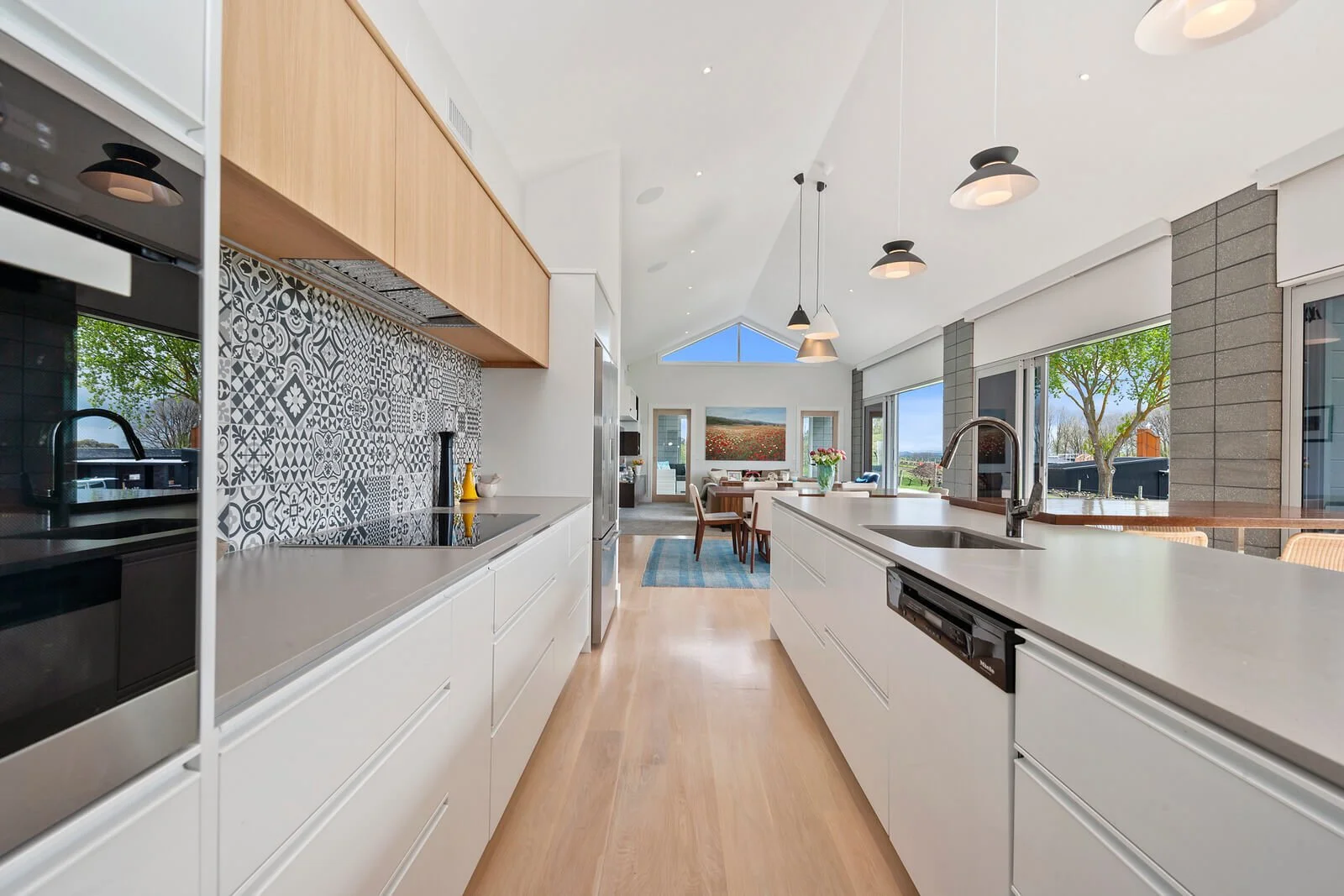 Modern kitchen with white cabinets, black appliances, patterned backsplash, wood accents, and a view into a dining and living area with large windows, trees outside, and contemporary lighting.