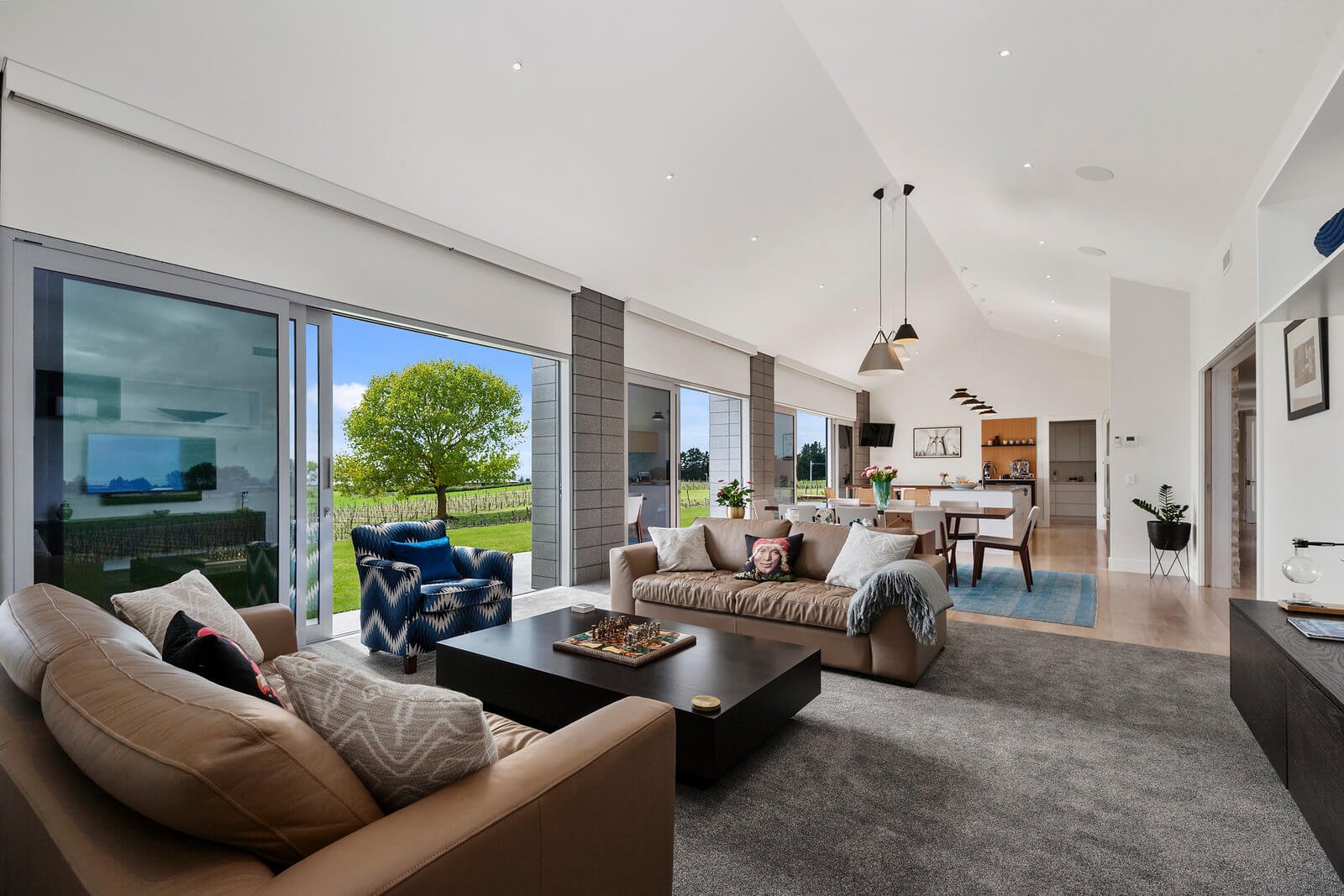 Spacious living room with beige sofas, a black coffee table, and large windows showing a green tree outside.