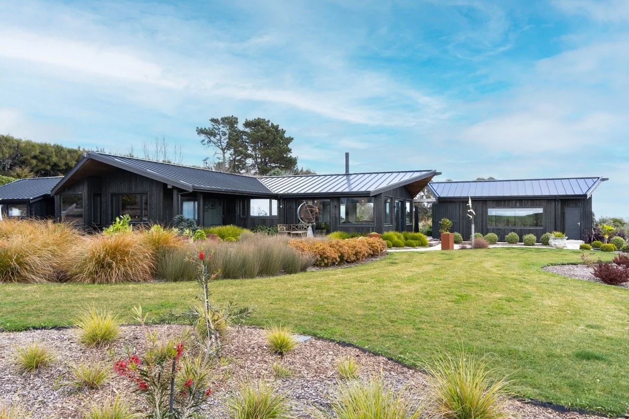Modern black house with metal roof, surrounded by landscaped yard and garden, under a blue sky.